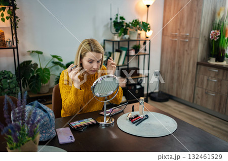 Woman applying makeup in a cozy indoor setting with plants and warm lighting during afternoon hours 132461529