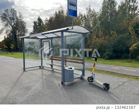 Modern bus stop with transparent shelter, wooden bench, and electric scooter parked nearby, surrounded by greenery and a paved pathway, illustrating urban transportation options Modern bus stop with transparent shelter, wooden bench, and electric scooter parked nearby, surrounded by greenery and a paved pathway, illustrating urban transportation options 132462167