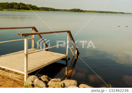 Tranquil lakeside scene featuring wooden dock extending over calm water, surrounded by lush greenery and distant trees, creating a serene natural atmosphere for relaxation 132462236
