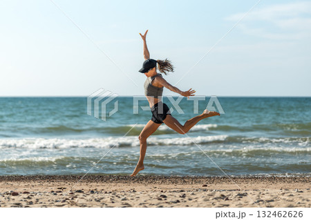 Dynamic female athlete on sunny shoreline, Cheerful woman in energetic motion leaping at coast with windblown hair 132462626