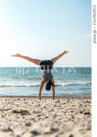 Joyful female athlete performing acrobatic move at coast, Playful lady showcasing athleticism with cartwheel by ocean 132462651
