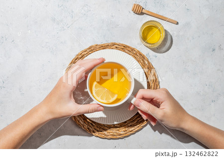 Hand holding cup of hot tea with lemon and honey on a white textured background with shadows. Hand holding cup of hot tea with lemon and honey on a white textured background with shadows. 132462822
