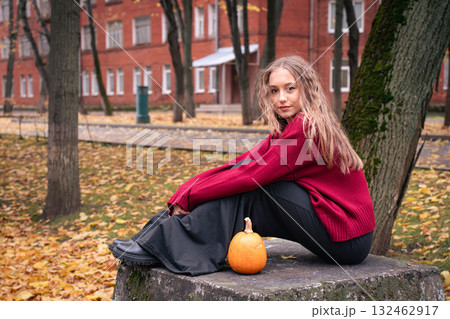 Teen girl in a red sweater sits curled up beside a pumpkin on a stone platform in an autumn park near Timiryazev Academy Teen girl in a red sweater sits curled up beside a pumpkin on a stone platform in an autumn park near Timiryazev Academy 132462917