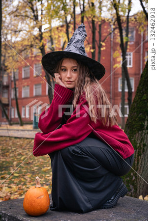 Teen girl in a witch hat and red sweater crouches beside a pumpkin in a park near Timiryazev Academy, surrounded by golden leaves and brick buildings 132462918