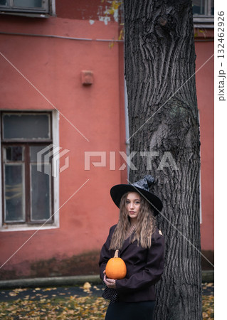 Teen girl in witch hat holding a pumpkin near a tree, surrounded by autumn leaves and soft natural light, creating a mysterious Halloween mood Teen girl in witch hat holding a pumpkin near a tree, surrounded by autumn leaves and soft natural light, creating a mysterious Halloween mood 132462926