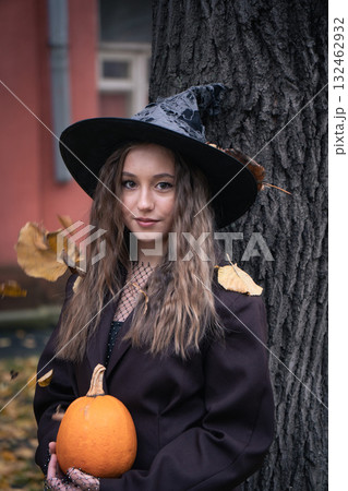 Teen girl in witch hat holding a pumpkin near a tree, surrounded by autumn leaves and soft natural light, creating a mysterious Halloween mood 132462932