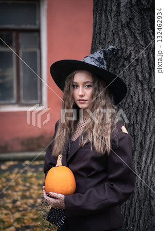 Teen girl in witch hat holding a pumpkin near a tree, surrounded by autumn leaves and soft natural light, creating a mysterious Halloween mood 132462934