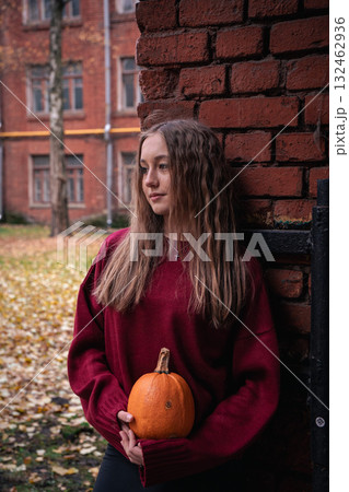 Teen girl in a red sweater holds a pumpkin while leaning against an old brick wall with autumn leaves on the ground in the Timiryazevs Academy, Moscow 132462936