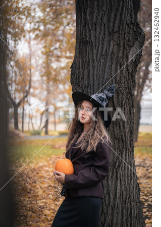 Teen girl in witch hat holding a pumpkin near a tree, surrounded by autumn leaves and soft natural light, creating a mysterious Halloween mood 132462940