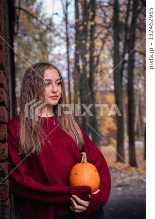 Teen girl in a red sweater holds a pumpkin while leaning against an old brick wall with autumn leaves on the ground in the Timiryazevs Academy, Moscow 132462955