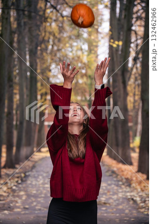 Teen girl in a red sweater throws a pumpkin into the air in an autumn park, surrounded by tall trees and fallen leaves Teen girl in a red sweater throws a pumpkin into the air in an autumn park, surrounded by tall trees and fallen leaves 132462966
