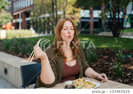 Plus size woman having lunch outdoors, eating noodles. 132463029