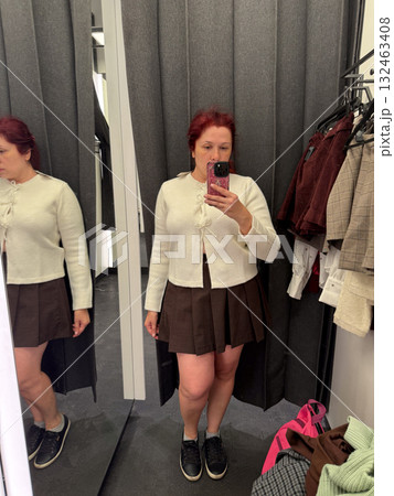 Woman trying on cream cardigan and brown skirt in fitting room. Modern fashion, mirror reflection, and everyday shopping experience. 132463408