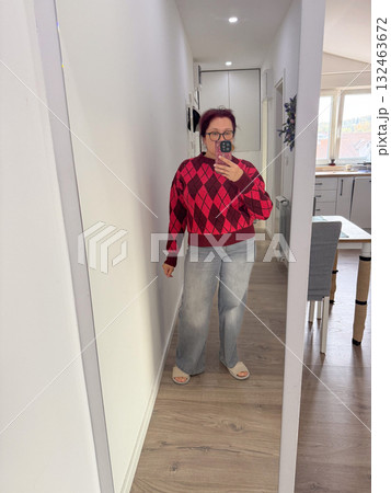 Woman in red argyle sweater and gray jeans taking mirror selfie. Casual outfit, cozy atmosphere, and modern home lifestyle. 132463672