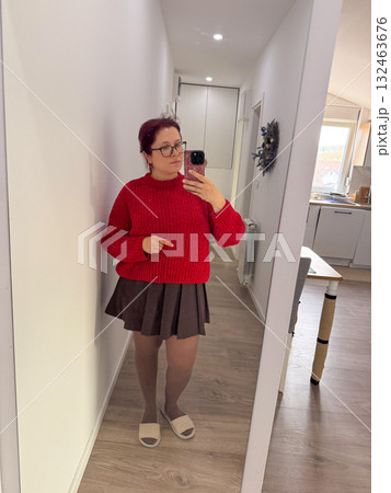 Woman in red knitted sweater and brown skirt taking mirror selfie. Bright accent, cozy mood, and daily outfit inspiration. 132463676