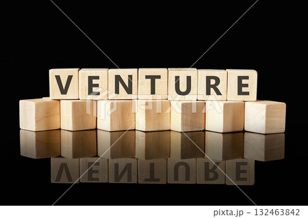 A stack of light brown wooden blocks spells the word VENTURE on a glossy black surface. The arrangement reflects the word, symbolizing innovation and ambition 132463842