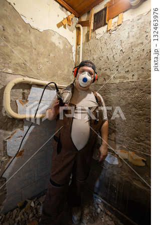 A man stands in a room with bare walls, a drill slung over his shoulder. A worker holds the drill near a wall of exposed plaster and concrete, preparing to make repairs. 132463976