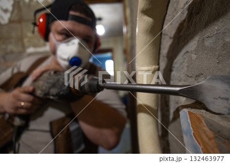 A construction worker using a hammer drill to remove old plaster from an interior wall during a home renovation is performing demolition work while wearing personal protective equipment. 132463977