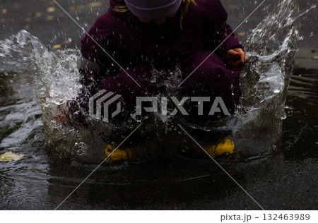 A small child in yellow boots and a purple jumpsuit joyfully jumps and splashes in a puddle. He jumps into the puddle, creating a huge splash. A carefree childhood in the fresh air. 132463989