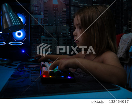 Young child focusing intently on a computer screen, hands on a colorful backlit keyboard and mouse, engaging with technology in a dim room with a futuristic cityscape background. 132464017