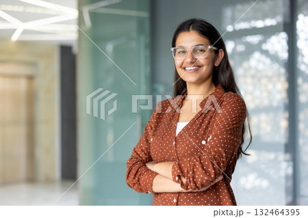 Successful confident businesswoman at workplace inside office, woman with crossed arms smiling and looking at camera, standing near window. Satisfied office worker of company 132464395
