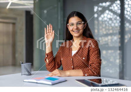 Young indian business woman sitting at a desk and raising her hand, smiling while participating in a video call or online meeting in a modern office environment Young indian business woman sitting at a desk and raising her hand, smiling while participating in a video call or online meeting in a modern office environment 132464416
