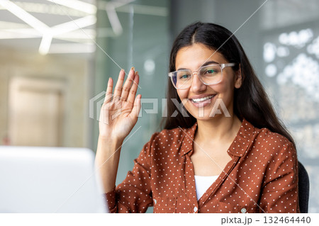 Happy young businesswoman wearing glasses, waving at a laptop screen during an online meeting, engaging in distant negotiations from a modern office Happy young businesswoman wearing glasses, waving at a laptop screen during an online meeting, engaging in distant negotiations from a modern office 132464440