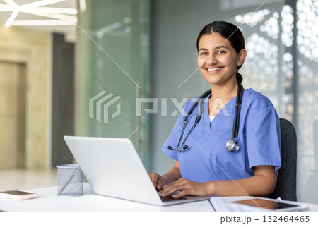 Confident and smiling, a young nurse in blue scrubs and a stethoscope works diligently on her laptop in a bright, modern hospital environment, reflecting professionalism and dedication 132464465
