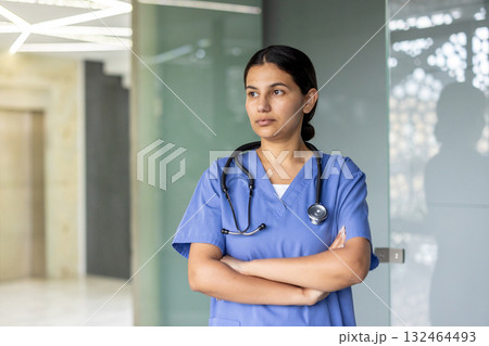 Healthcare professional woman wearing blue scrubs and a stethoscope, standing with arms crossed and looking away, representing dedication and critical thinking in her medical role Healthcare professional woman wearing blue scrubs and a stethoscope, standing with arms crossed and looking away, representing dedication and critical thinking in her medical role 132464493