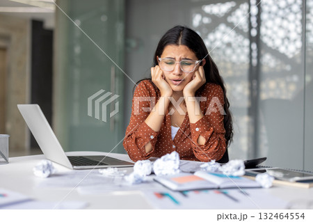 Young woman feeling overwhelmed and frustrated by her work, sitting at a desk with a laptop and crumpled papers, showing signs of stress and a tight deadline 132464554