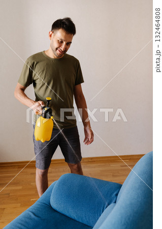 Happy smiling young man spraying water using yellow plastic sprayer on blue sofa in stylish living room, engaging in thorough home cleaning and maintenance. Concept of household cleanliness. 132464808