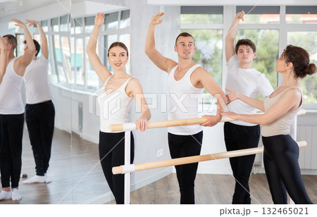 Professional female ballet trainer teaching group of dancers, helps to coordinate movements correctly. Young girl and guys near ballet barre in dancing hall 132465021