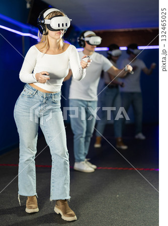 In darkened hall of gaming club, girl in VR helmet takes part in group game 132465025
