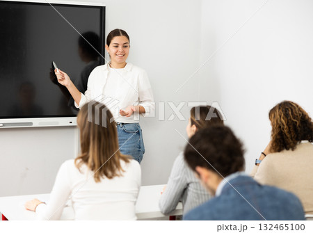 Young female teacher near interactive board, gesturing with hand in front of people sitting at desks in auditory 132465100