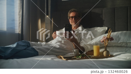 Mature Executive Sits on Hotel Bed, Dressed in Bathrobe, Checking Smartphone While Holding Coffee Mature Executive Sits on Hotel Bed, Dressed in Bathrobe, Checking Smartphone While Holding Coffee 132468026