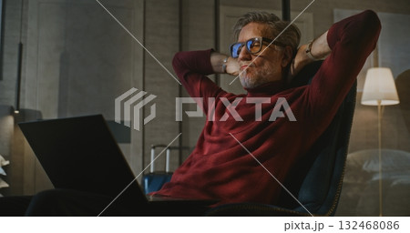 Gray Haired Man Sits by Bed, Working on Laptop in Evening Light of Hotel Room 132468086