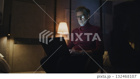 Mature Traveler Works on Laptop in Softly Lit Modern Hotel Room at Night 132468107