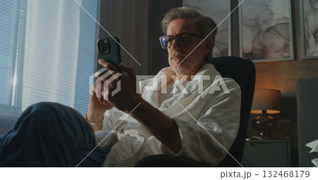 Gray Haired Man in Robe Sits by Window, Confirming Online Purchase With Phone and Credit Card Gray Haired Man in Robe Sits by Window, Confirming Online Purchase With Phone and Credit Card 132468179