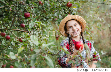 Cheerful girl stands on background an apple orchard with red apples, tastes, enjoys snacks, vitamins, organic fruits. She is dressed in checkered shirt and hat. Cheerful girl stands on background an apple orchard with red apples, tastes, enjoys snacks, vitamins, organic fruits. She is dressed in checkered shirt and hat. 132468510