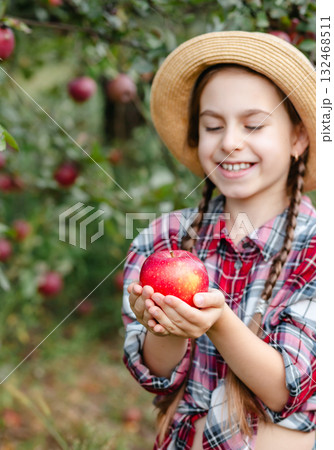 Cheerful girl stands on background an apple orchard with red apples, tastes, enjoys snacks, vitamins, organic fruits. She is dressed in checkered shirt and hat. Cheerful girl stands on background an apple orchard with red apples, tastes, enjoys snacks, vitamins, organic fruits. She is dressed in checkered shirt and hat. 132468511