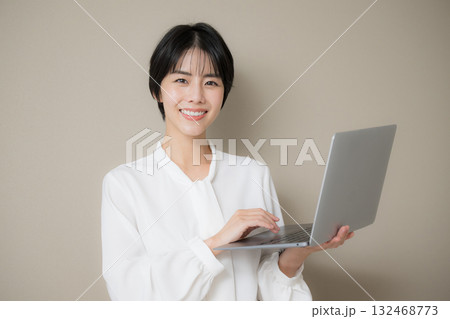 ベージュ背景でパソコンを持つポーズのかわいい黒髪の美人な女性カメラ目線 ベージュ背景でパソコンを持つポーズのかわいい黒髪の美人な女性カメラ目線 132468773