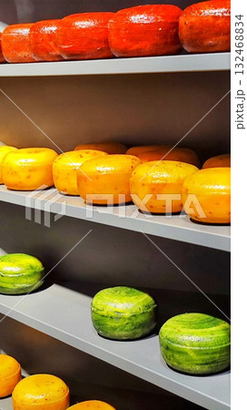 Assortment of cheeses displayed on gray shelves. Top shelf holds red wax-covered rounds, middle has orange rounds, and bottom features green wax-covered rounds. Netherlands, Holland cheese  132468834