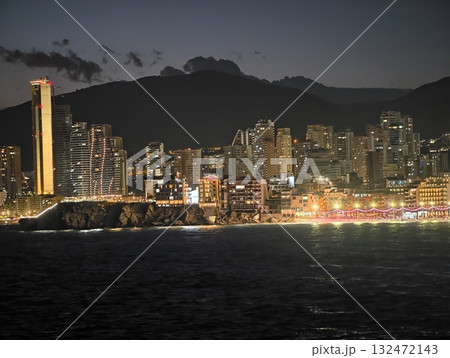 bright resort lights along coast, golden waves reflect festive lights beneath mountain silhouette 132472143