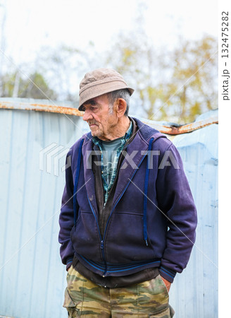 Elderly Russian man standing in a rural village, hands in pockets, looking sideways, dressed in casual clothes and hat. 132475282