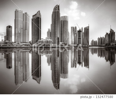 Monochrome city skyline with tall skyscrapers reflecting on calm water under a cloudy sky, Dubai Marina bay UAE 132477580