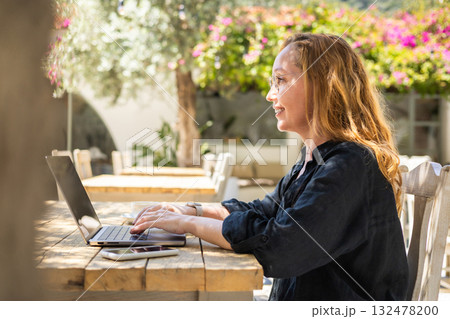 Side View of Smiling Woman Working on Laptop Outdoors in Cafe with Greenery and Natural Light 132478200