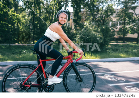 Happy active senior woman in helmet riding red bicycle on sunny path in green urban park 132478248