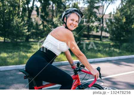 Confident mature woman smiling on red bicycle in activewear and helmet on sunny park bike lane Confident mature woman smiling on red bicycle in activewear and helmet on sunny park bike lane 132478256