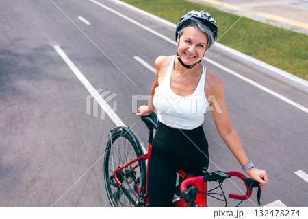 Happy Senior Woman in Helmet Standing with Bicycle on City Road in Sportswear and Smiling 132478274