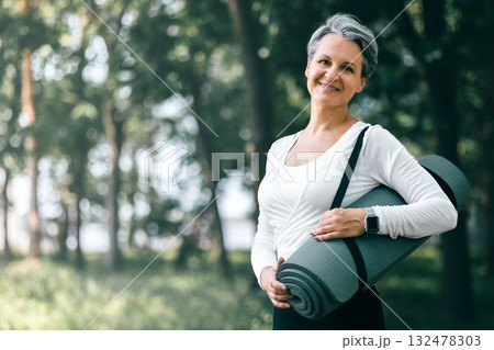 Smiling Mature Woman Holding Yoga Mat in Forest Park Before Outdoor Wellness Session 132478303